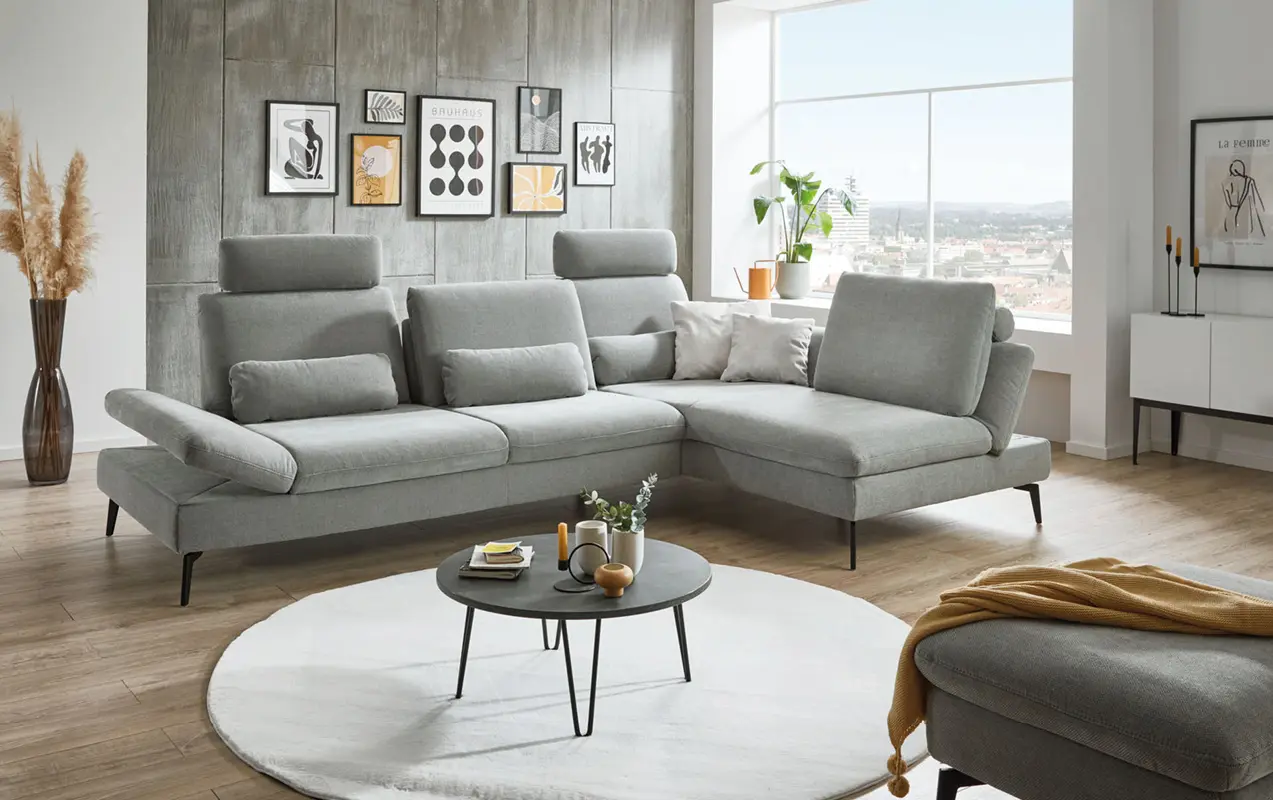 Graue Eckcouch in modernem Wohnzimmer mit Couchtisch und Wandbildern, viel Tageslicht durch große Fenster.